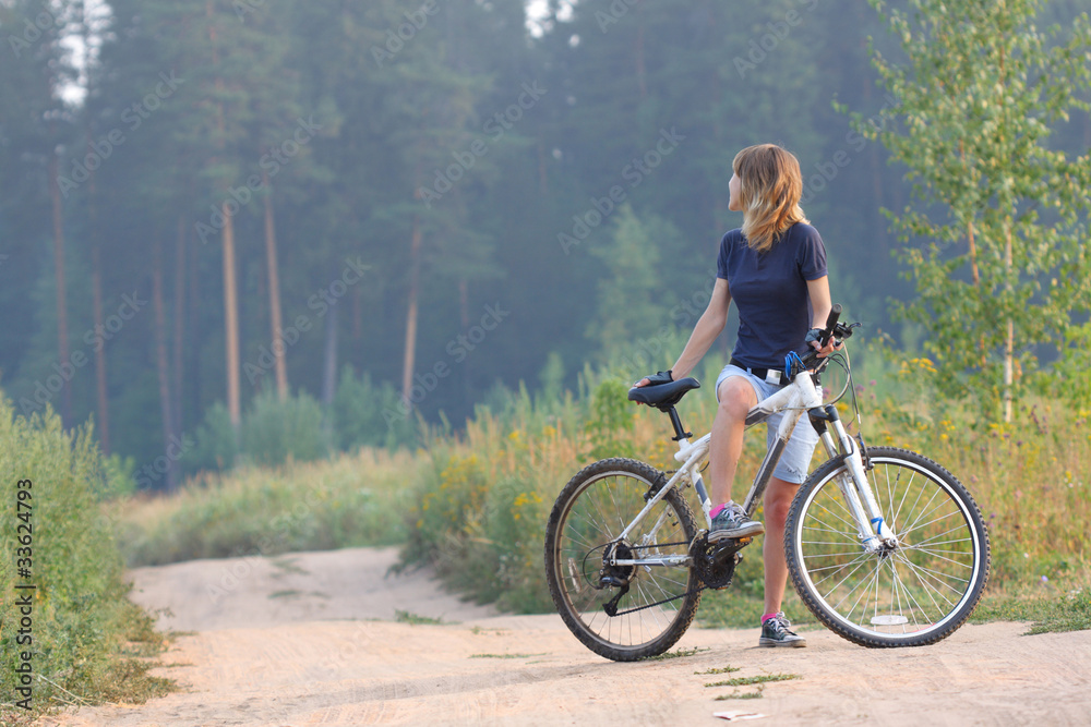 Young woman standing with a bicycle on a rural countryside road and looking back Young woman standing with a bicycle on a rural countryside road and looking back