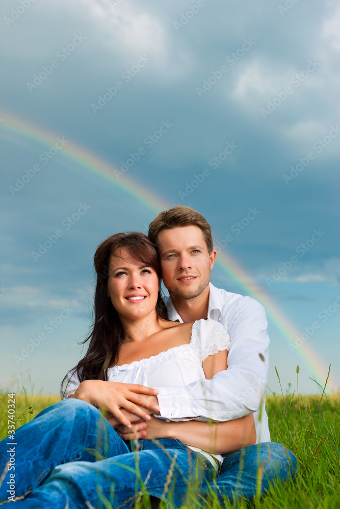 Happy couple sitting on a meadow under rainbow Happy couple sitting on a meadow under rainbow