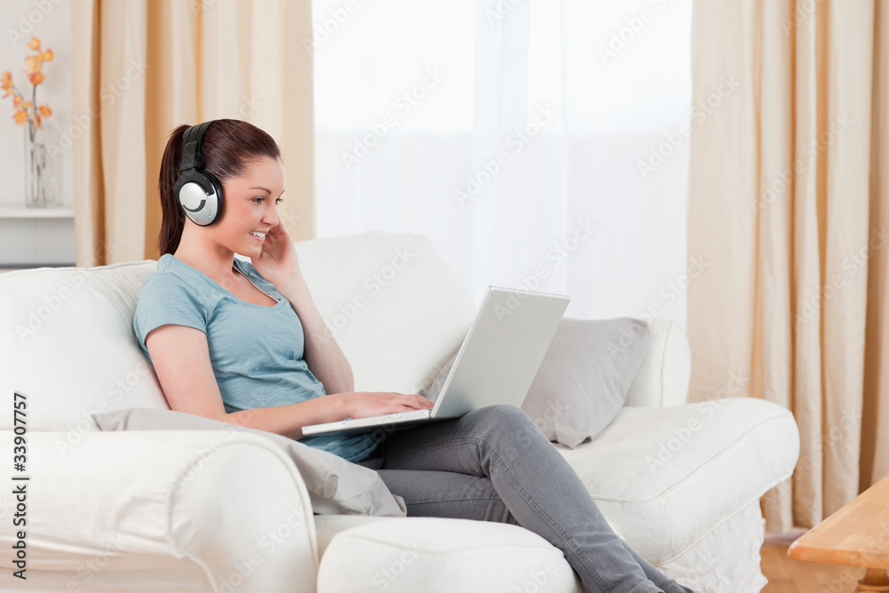 Gorgeous woman with headphones relaxing with her laptop while si Gorgeous woman with headphones relaxing with her laptop while si