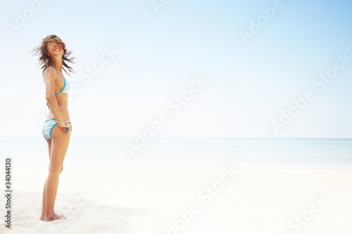 Young happy smiling woman in a swimsuit standing on a white-sand by the blue sea on clear blue sky background © Dudarev Mikhail
