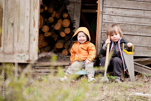 Two little children sit near a shed with garden tools