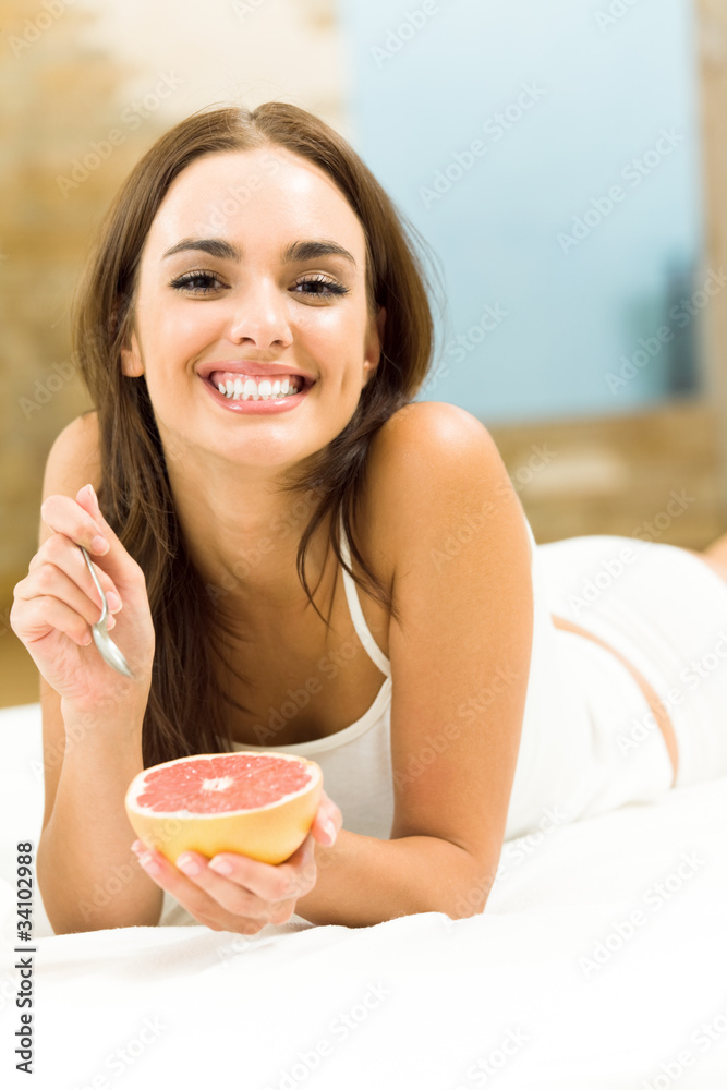 Smiling beautiful woman eating grapefruit on bed, at home
