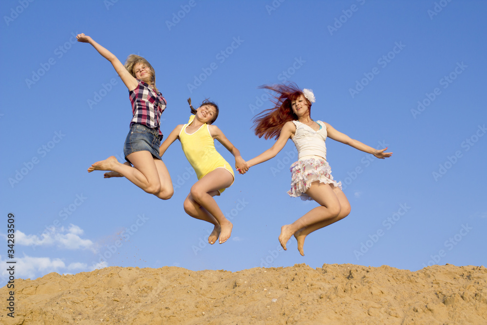 Three girls jump above sand embankment against blue sky
