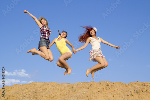 Three girls jump above sand embankment against blue sky