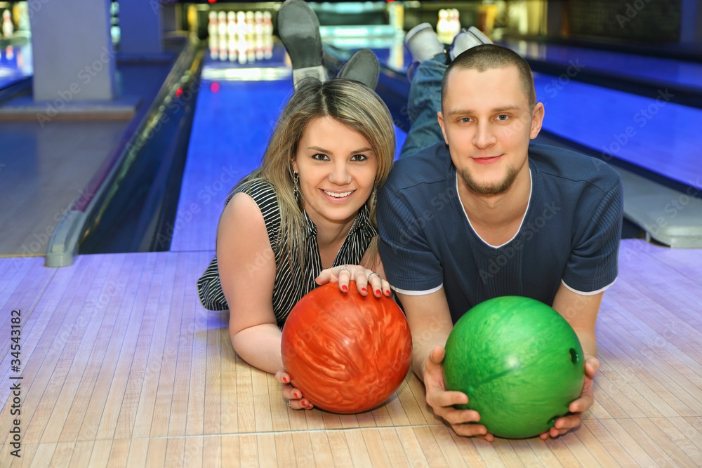 Fototapeta premium Girl and man lie on parquet in club and hold balls for bowling