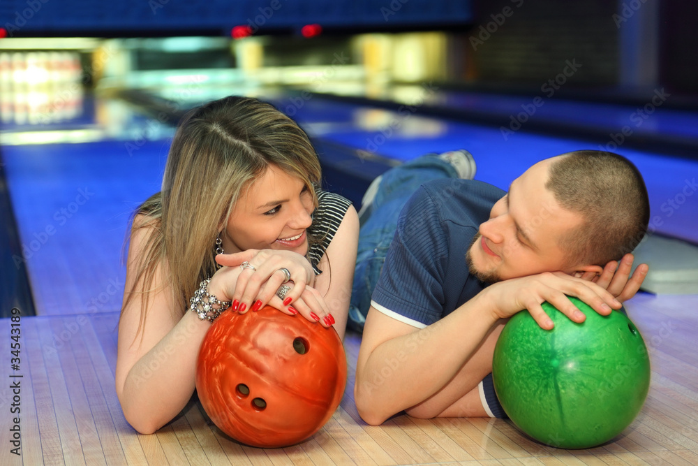 Fototapeta premium Couple lie on parquet, look on each other with balls for bowling