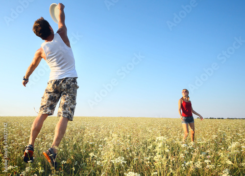 Young couple playing frisbee on a green meadow with grass on clear blue sky background. Focus on a woman, man is motion blurred