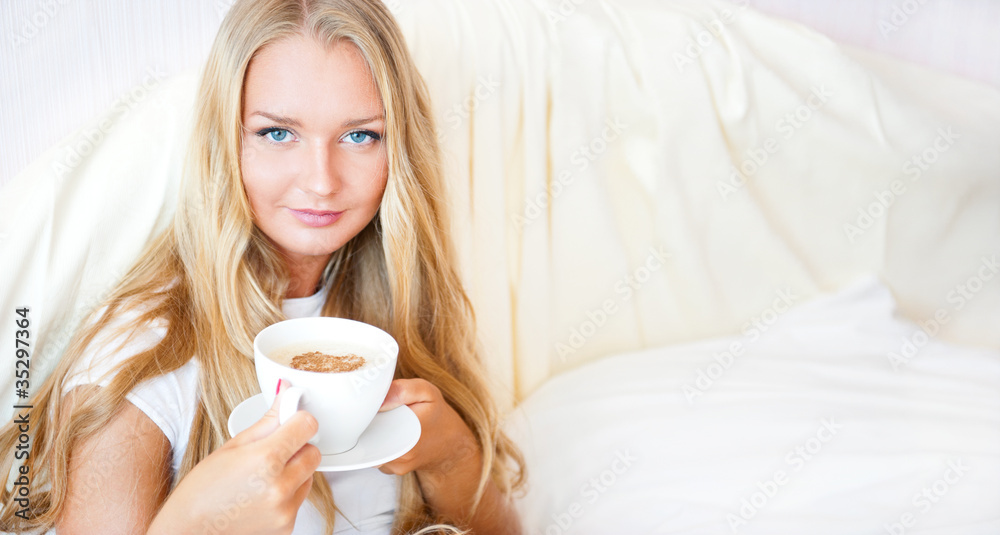 Smiling woman drinking a coffee lying on a bed at home or hotel.