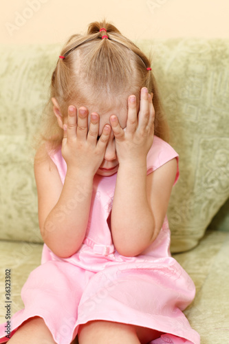 Little girl in pink dress on sofa closes face by hands