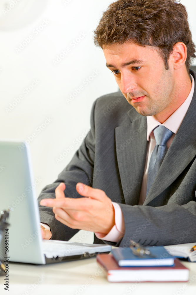 Confused businessman sitting at desk and working on laptop Confused businessman sitting at desk and working on laptop