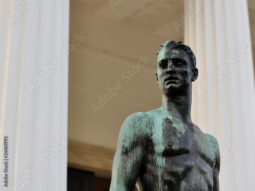 bronze statue of a athletic male body in front of a greek temple