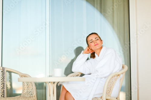 Woman in bathrobe sitting at table on terrace and relaxing