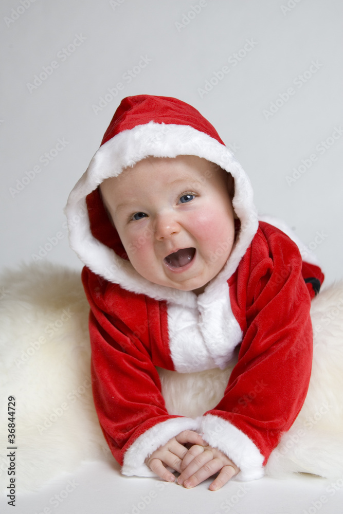 Kleines Baby als Weihnachtsmann verkleidet liegt lachend auf einem weißen Fell auf dem Bauch Kleines Baby als Weihnachtsmann verkleidet liegt lachend auf einem weißen Fell auf dem Bauch