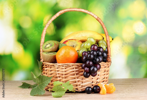 Fotografía Ripe juicy fruits in basket on wooden table on green background