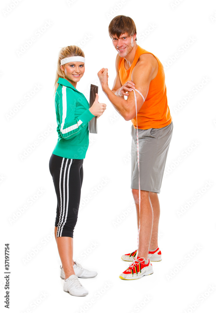 Male measuring biceps and girl in sportswear showing thumbs up Male measuring biceps and girl in sportswear showing thumbs up