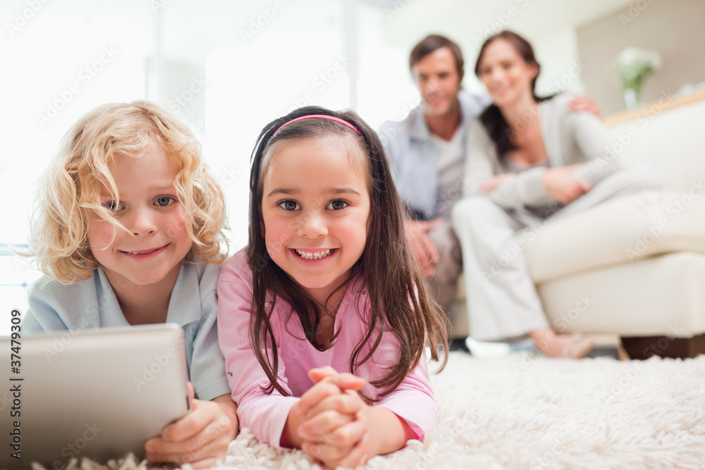 Siblings using a tablet computer while their parents are in the Siblings using a tablet computer while their parents are in the