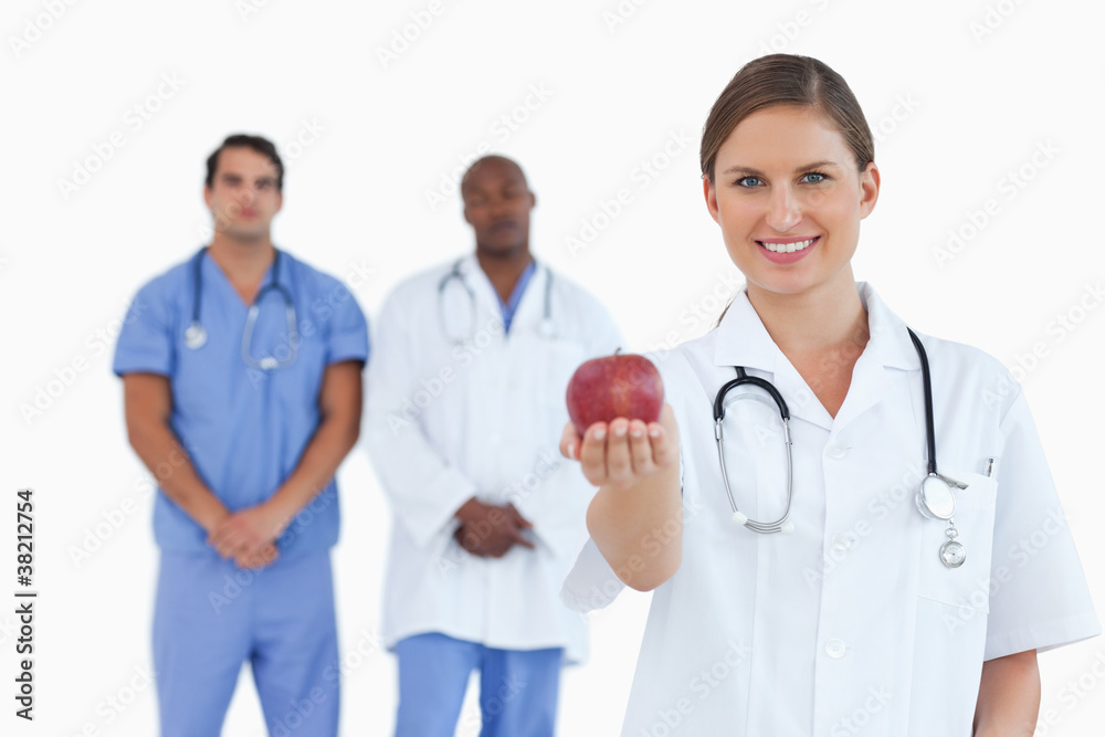 Doctor offering apple with colleagues behind her Doctor offering apple with colleagues behind her