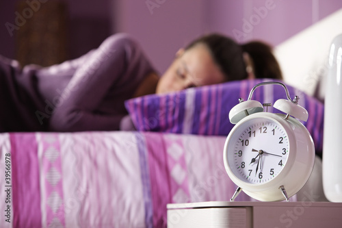 An alarm clock with a sleeping young woman in the background