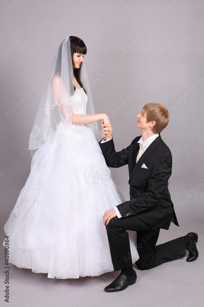 Happy groom kneels and holds beautiful bride hand in studio