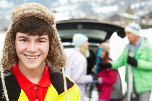 Teenage Boy Smiling At Camera Whilst Family Load Skis In Boot Of