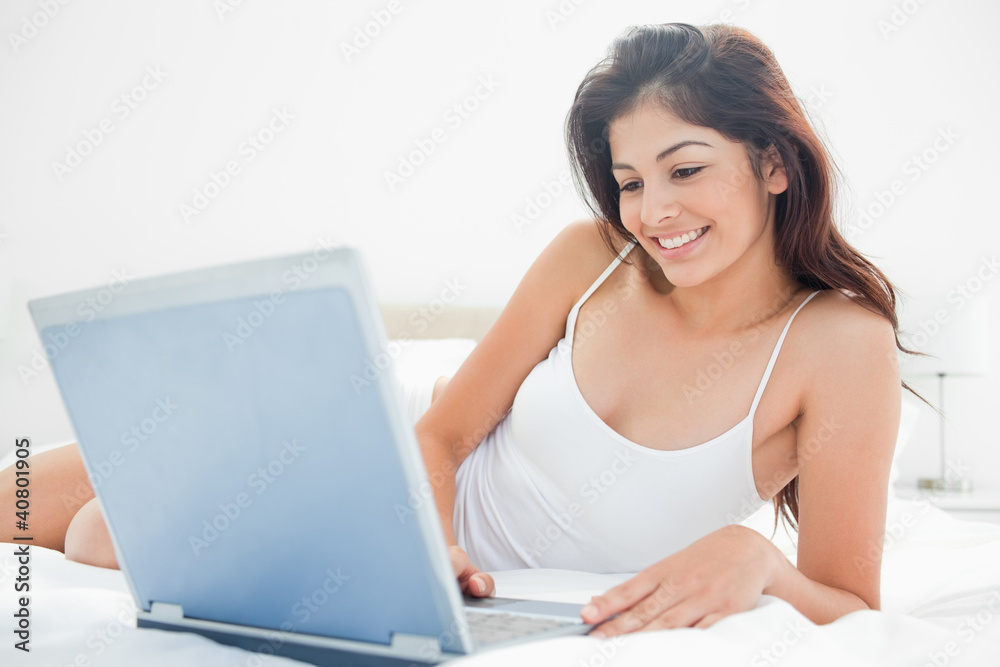 Woman relaxing across the bed with her laptop, enjoying what she Woman relaxing across the bed with her laptop, enjoying what she