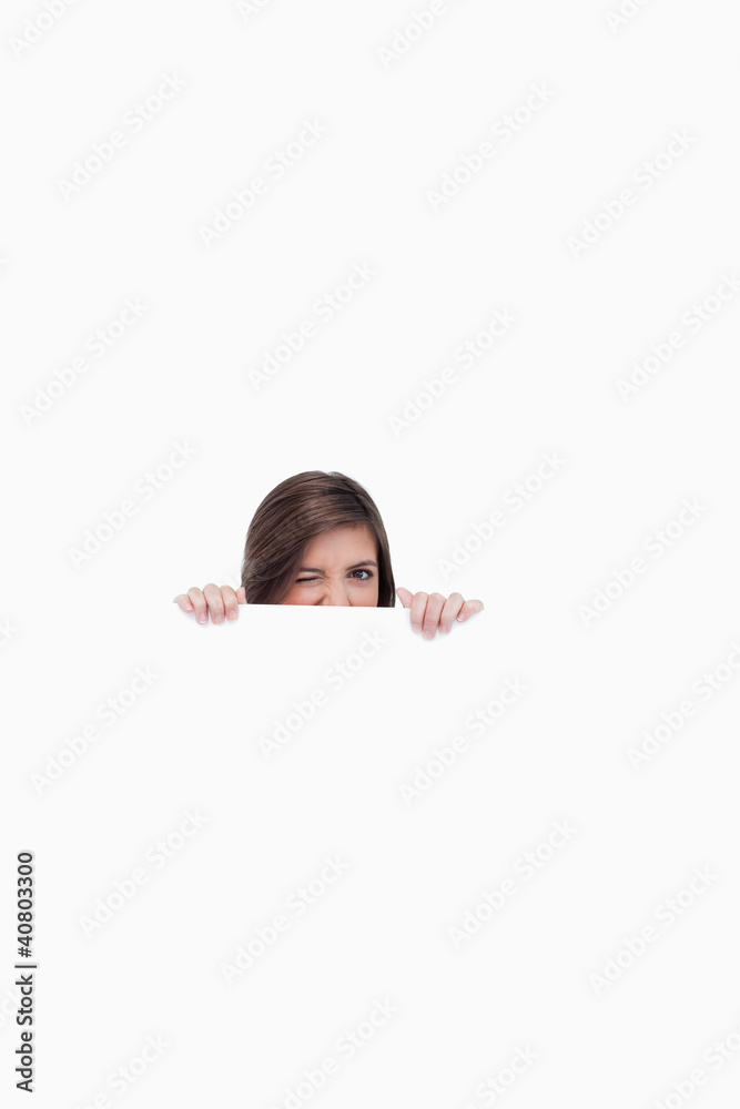 Young woman hiding behind a blank poster while showing a wink of Young woman hiding behind a blank poster while showing a wink of