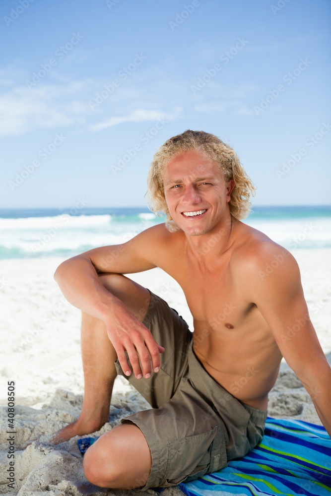 Smiling man sitting on his beach towel while looking at the came Smiling man sitting on his beach towel while looking at the came
