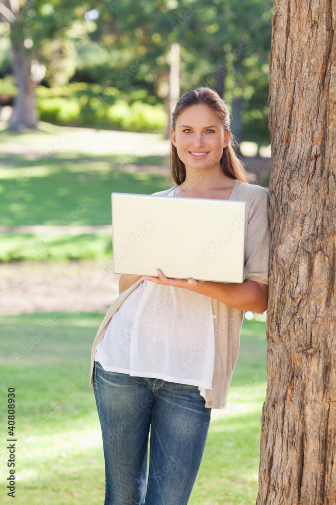 Smiling woman using a laptop while leaning against a tree Smiling woman using a laptop while leaning against a tree