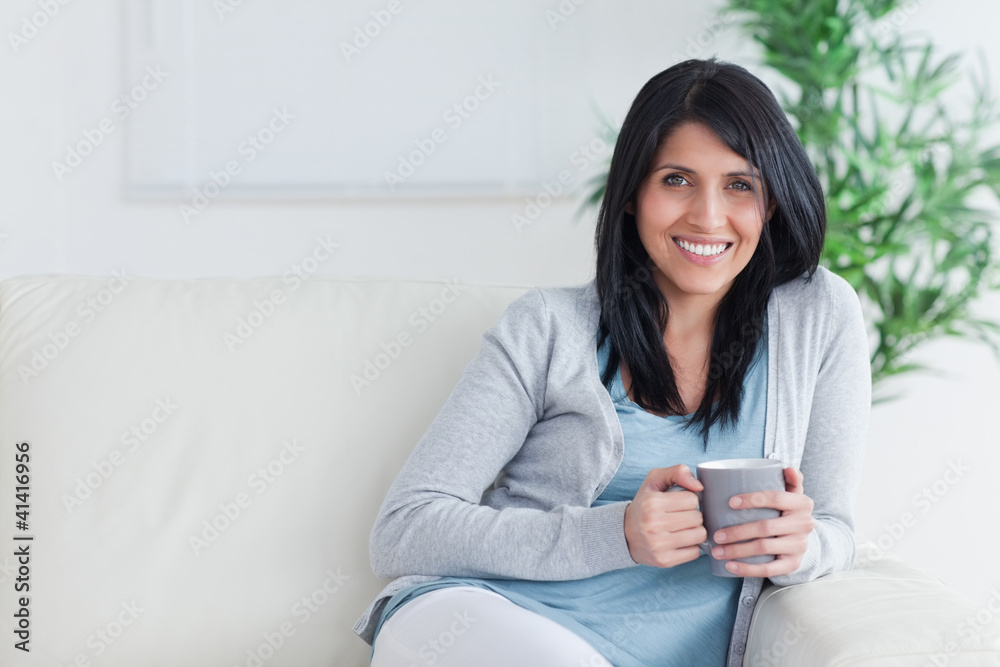 Smiling woman holding a mug with two hands while resting on a so Smiling woman holding a mug with two hands while resting on a so