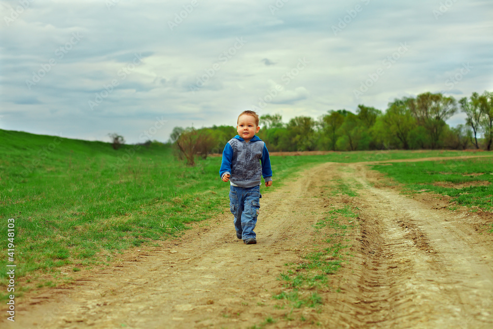 little baby boy walking country road on cloudy sky background