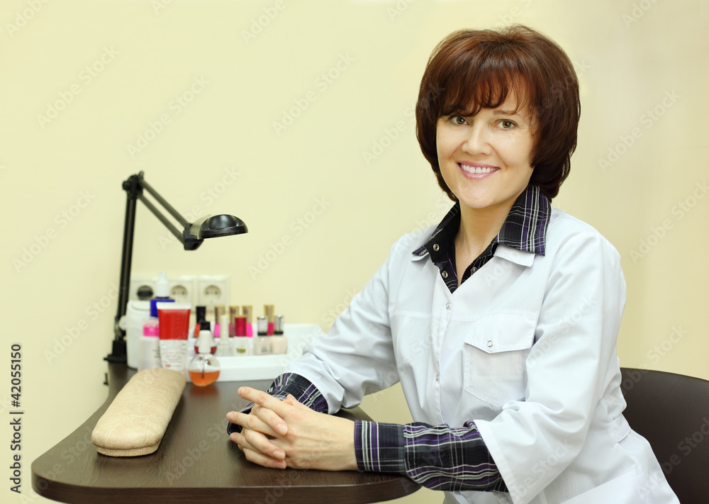 Smiling manicurist dressed in robe sits at table for manicure Smiling manicurist dressed in robe sits at table for manicure