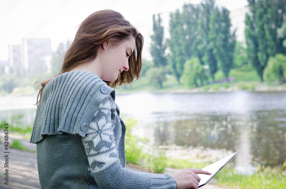 Fototapeta premium Girl sitting outdoors using a touchscreen tablet along a riverba