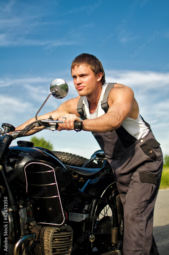 Handsome man in a boilersuit rolling a motorcycle.