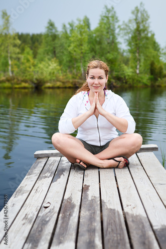 Happy woman in lotus pose sitting on wooden boards on river edge