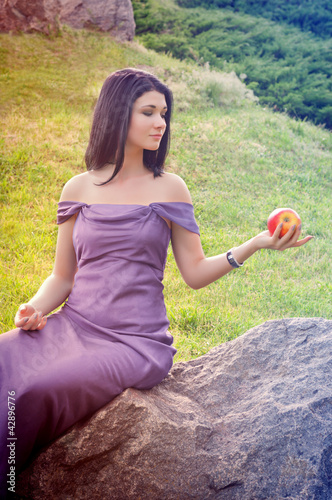 Young woman in dress sitting on a rock and holds an apple in his