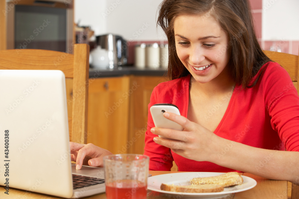 Teenage Girl Using Laptop And Mobile Phone Whilst Eating Breakfa