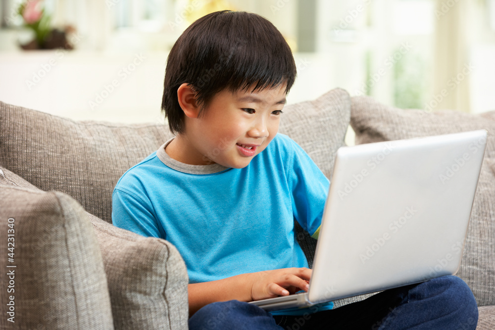 Young Chinese Boy Using Laptop Whilst Sitting On Sofa At Home Young Chinese Boy Using Laptop Whilst Sitting On Sofa At Home