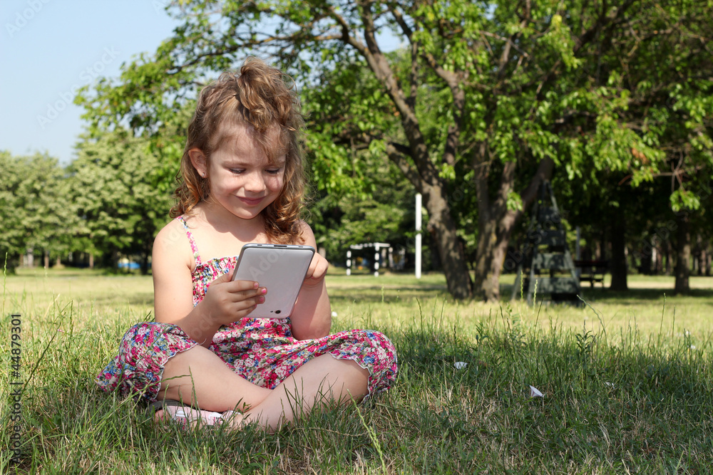 Fototapeta premium little girl sitting on grass and play with tablet pc