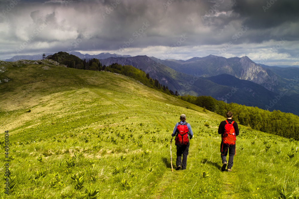 Obraz premium Two hikers on green meadow surrounded by high mountains