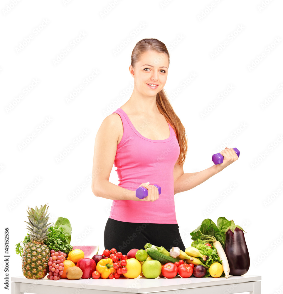 Smiling woman holding weights and table full of fruits and veget Smiling woman holding weights and table full of fruits and veget