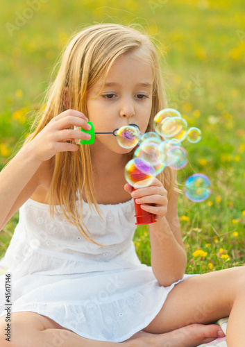 Cute little girl blowing soap bubbles and sitting on the meadow