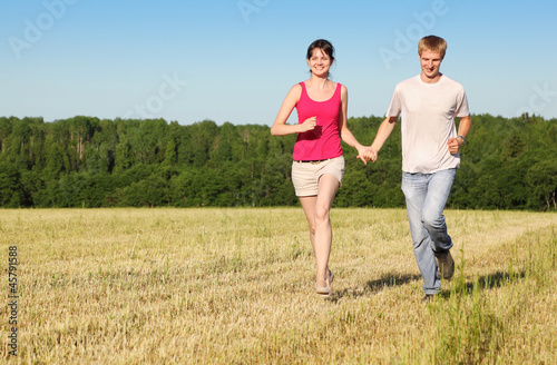 Husband, wife holding hands run in field near wood