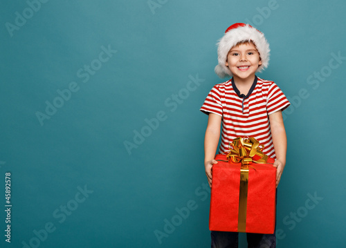 Funny child in Santa red hat holding Christmas gift in hand.