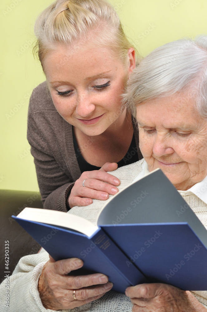 Senior woman with her caregiver in home reading book.