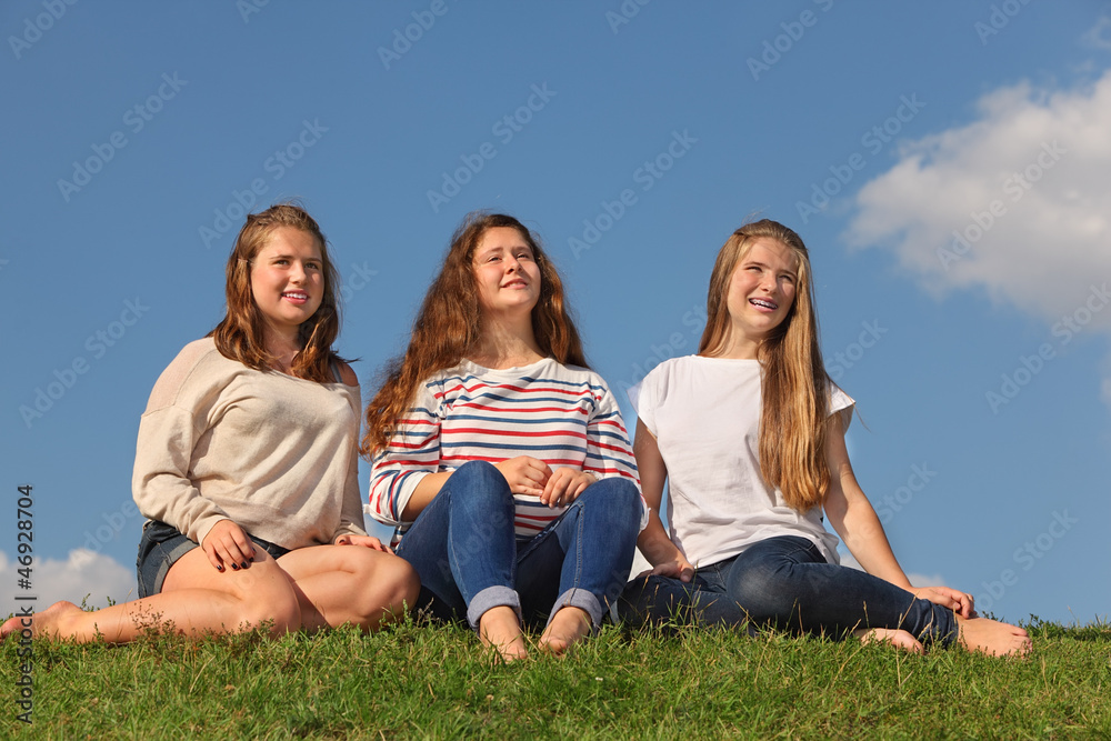 Three barefoot girls sit at green grass and look into distance