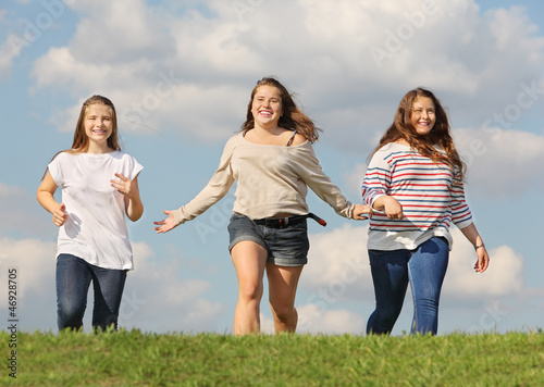 Fotografia Three girls run at green grass and smile at background of sky