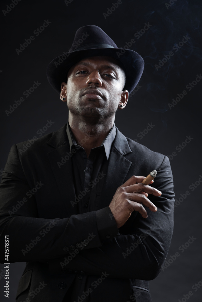 Black man wearing suit and hat gangster style smoking cigar. Black man wearing suit and hat gangster style smoking cigar.