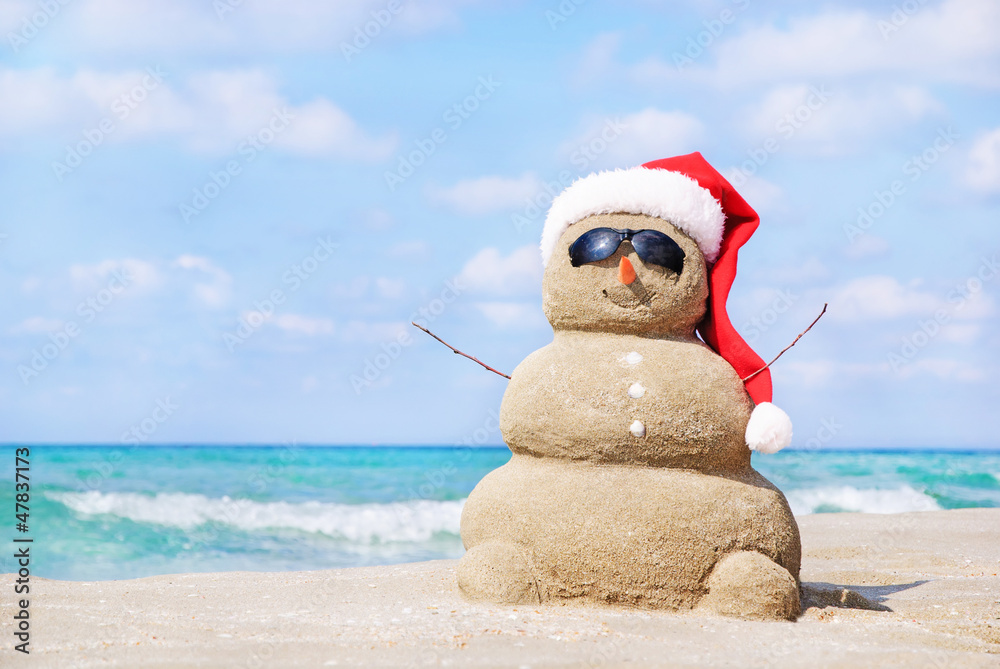 Smiling sandy snowman in red santa hat on the sea beach. - obrazy, fototapety, plakaty 