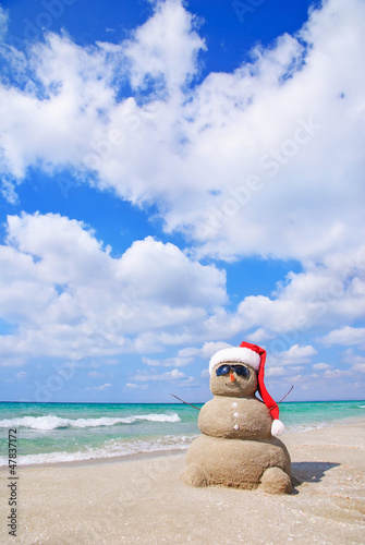 Smiling sandy snowman in red santa hat on the sea beach.