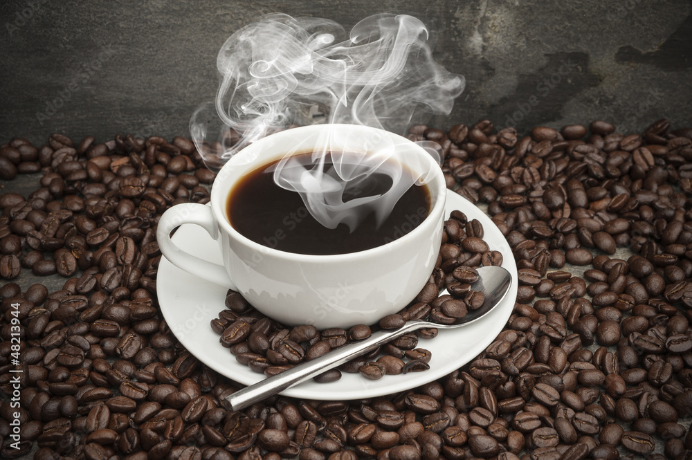 Fototapeta premium Steaming hot cup of coffee surrounded by dark coffee beans with